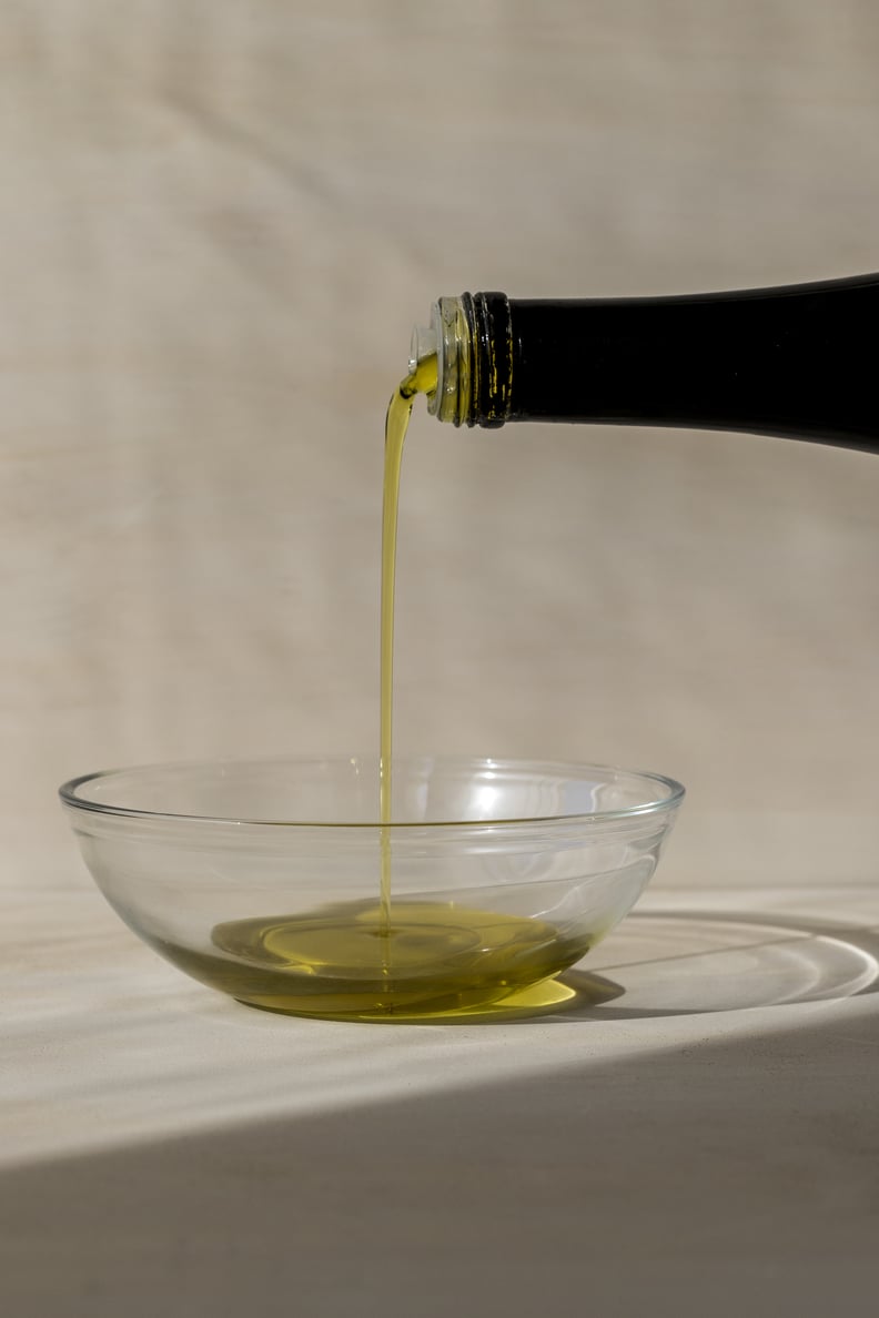 Image of the top of a black olive oil bottle with the contents being poured into a clear glass bowl. A side view photographed in natural light against a neutral background.